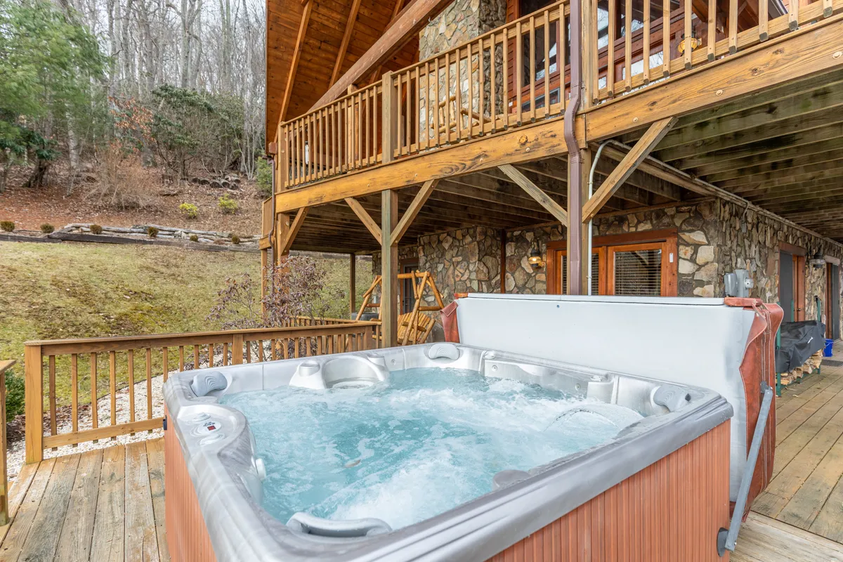 A luxurious hot tub sits on a spacious wooden deck next to a rustic stone façade, with a balcony above and surrounded by lush greenery. The scene radiates tranquility, offering views ideal for relaxing at sunset or under a starlit sky.
