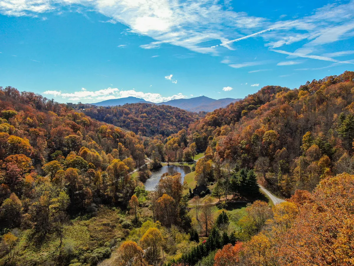 A sweeping vista of the Valle Crucis mountains featuring a vibrant tapestry of autumn foliage, a gently winding river, and layers of distant peaks, all beneath a sky dotted with soft clouds.