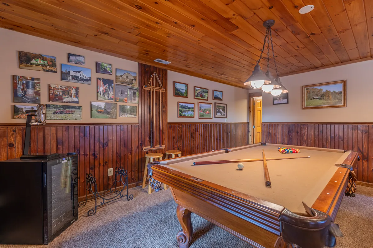 A casual and friendly game room setting with a prominently featured pool table, wooden walls lined with framed pictures, and attractive light fixtures overhead. The space encourages socializing, playing pool, and hosting gatherings, emphasizing the entertainment focus of the downstairs den.