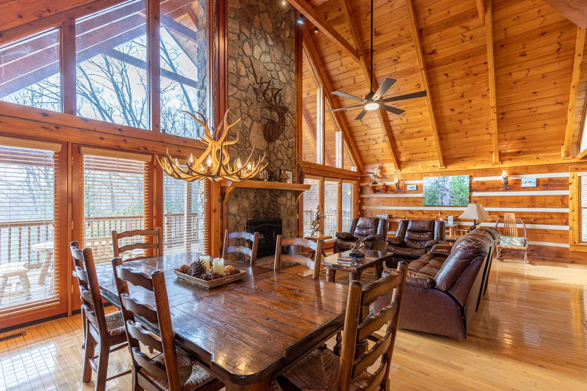 A spacious family and dining area featuring a dramatic stone fireplace, leather sofas, a large wooden dining table, and expansive windows framing views of the mountains. The space is bathed in natural light, accentuated by a rustic chandelier overhead.