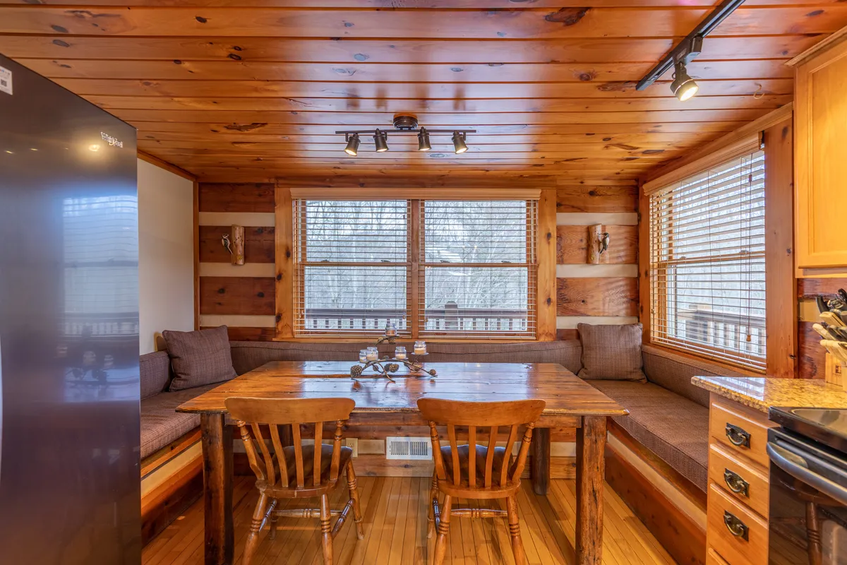 A sunlit dining area with a large wooden table, surrounded by comfortable seating and large windows that allow natural light to stream in. The adjacent kitchen area and view through the windows emphasize a close connection to the outdoors and the changing seasons.