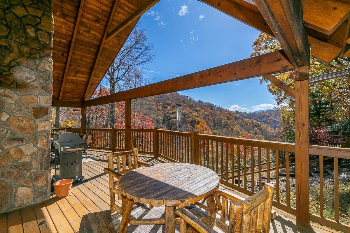 A rustic wooden deck furnished with a dining table and comfortable seating overlooks sweeping mountain views. The deck features a grill for barbecues and is surrounded by lush greenery, creating a serene space for outdoor dining, relaxation, and enjoying nature.