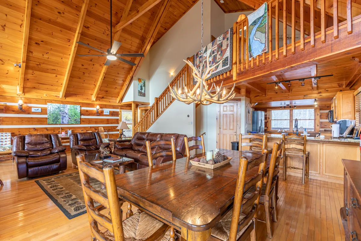 A wide-angle view of the open-concept living space showing the wooden beams, an antler chandelier above the dining table, leather sofas near the fireplace, and the adjacent kitchen area. Large windows offer glimpses of the outdoors, enhancing the warm, inviting atmosphere.