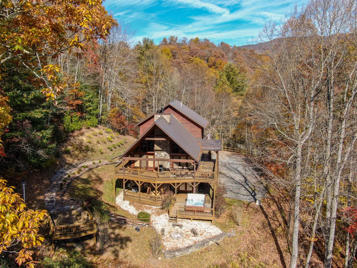 A welcoming cabin exterior with a wooden balcony and stone façade, nestled among mature trees and featuring a driveway that invites further exploration. The porch area hints at relaxing evenings or gatherings around a firepit under the stars.