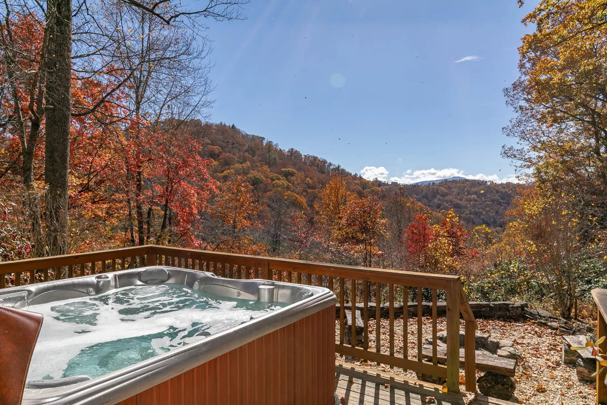 A bubbling hot tub set on a wooden deck faces a spectacular mountain vista with vibrant autumn foliage. The setting invites guests to unwind, socialize, and take in the tranquil natural surroundings, day or night.