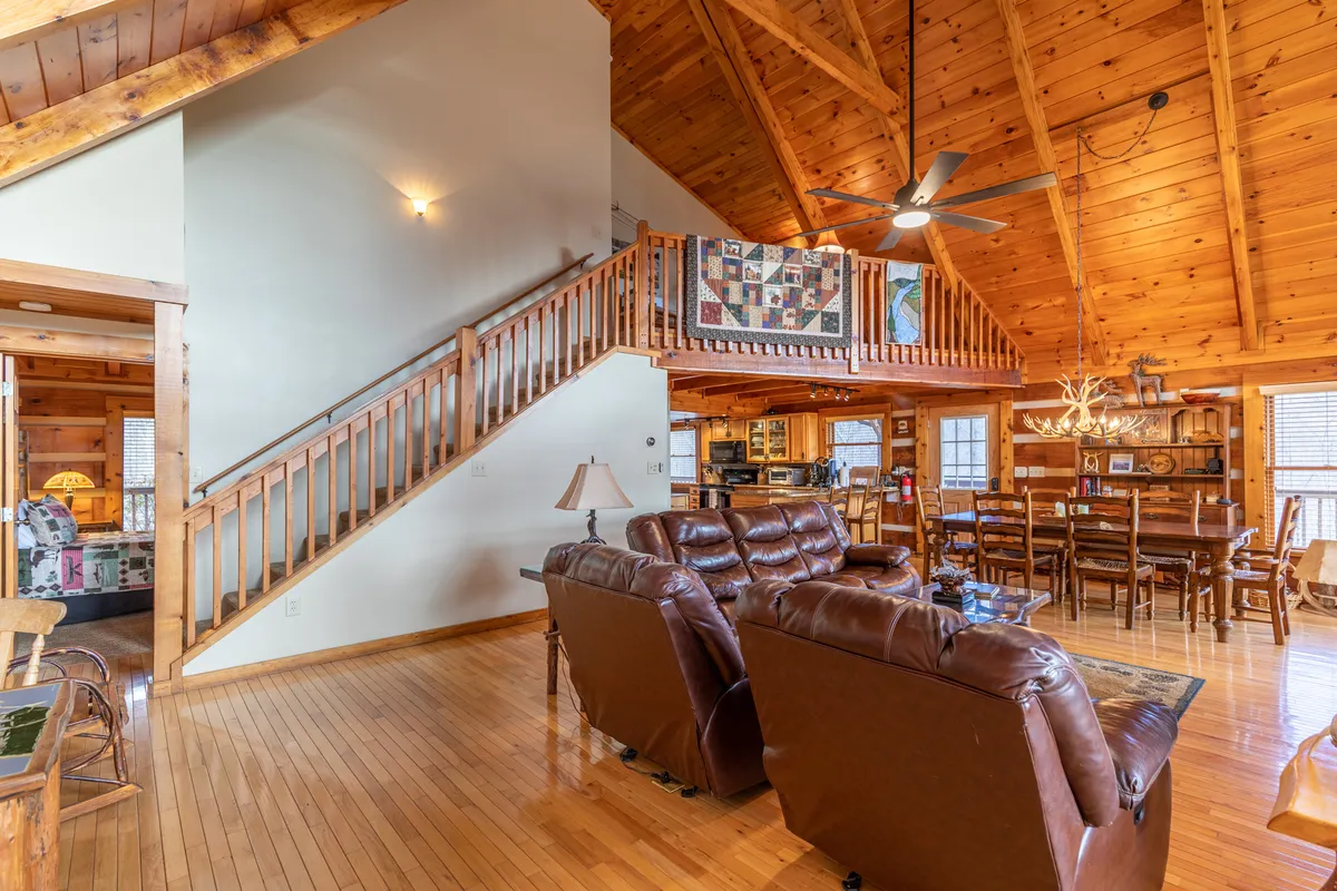 A spacious and inviting living area featuring a wooden staircase, leather sofas, a large dining table, and a quilt hanging on the railing. The exposed wooden beams and open layout create a sense of connection and warmth, with ample space for family gatherings and activities.