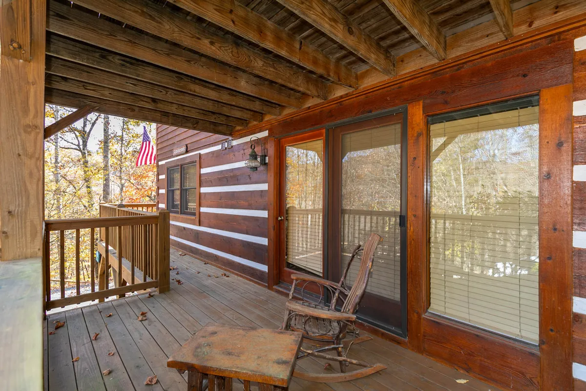 A cozy porch with a rocking chair, wooden railing, and a flag gently waving. Sliding glass doors connect the porch to the interior, while the view beyond features lush greenery, suggesting proximity to the outdoors and opportunities to enjoy nature.
