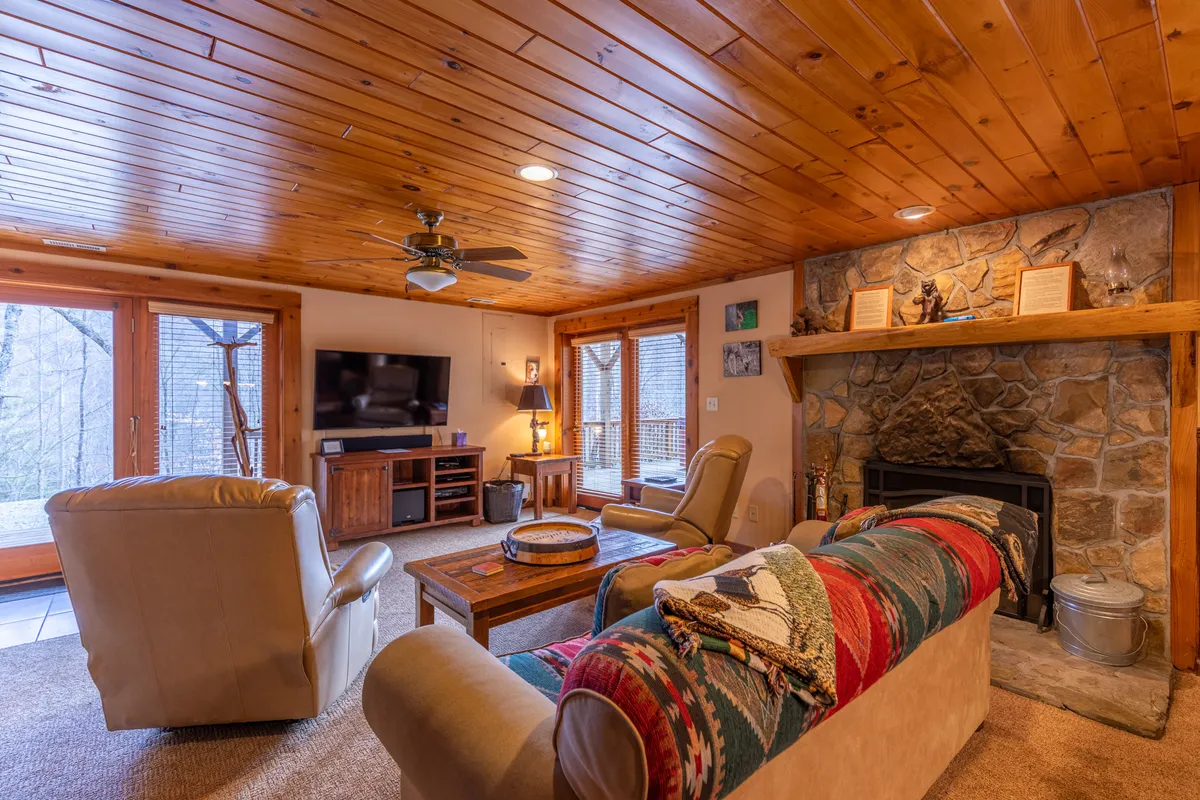 A comfortable seating area anchored by a stone fireplace and facing a large TV, with rustic wooden ceilings and big windows letting in gentle light. The layout invites relaxation, movie watching, and cozy evenings after active game sessions.