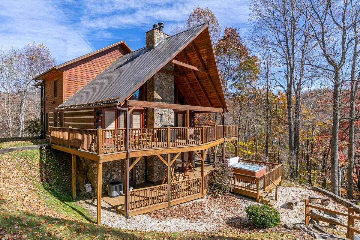 The image features the rustic stone and wood cabin exterior set in a lush grove of trees, with a spacious wooden deck showcasing a bubbling hot tub. The scene exudes warmth and tranquility, inviting guests to relax outdoors, dine al fresco, and soak in panoramic sunset views over Grandfather Mountain.