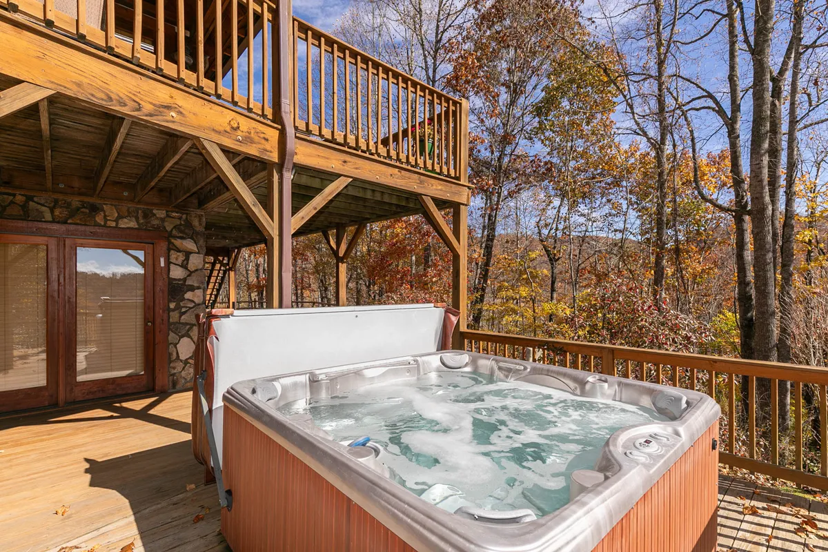 An exterior shot of a spacious wooden deck with a prominent hot tub, surrounded by lush trees. The scene suggests opportunities for stargazing, outdoor gatherings, and peaceful moments immersed in nature.