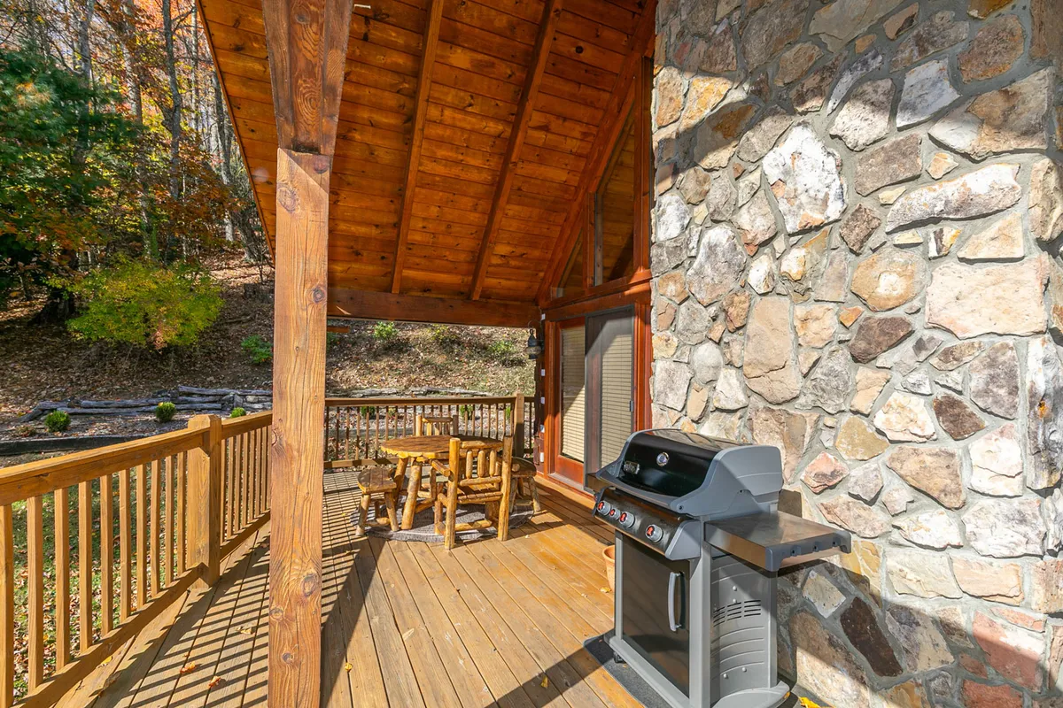 A wooden deck features a grill, wooden table, and sturdy railing overlooking the outdoors. The natural setting is visible beyond the deck, suggesting an ideal spot for barbecuing, casual meals, or stargazing after a day by the lake.