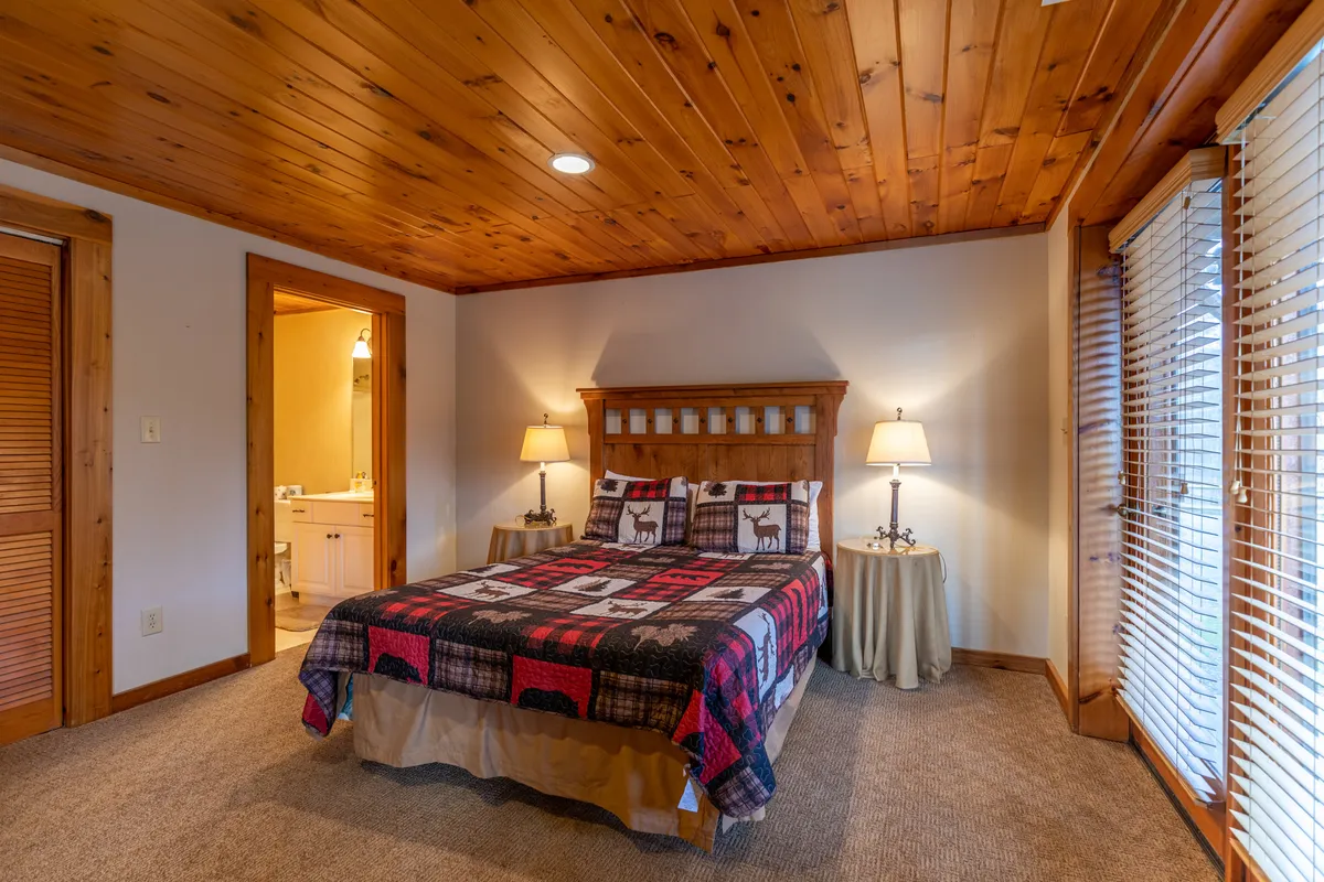 A comfortable bedroom with a bed adorned in a patterned quilt, nightstands with warm glowing lamps, and a wooden ceiling. The space is bathed in gentle natural light, creating a peaceful, restful atmosphere.