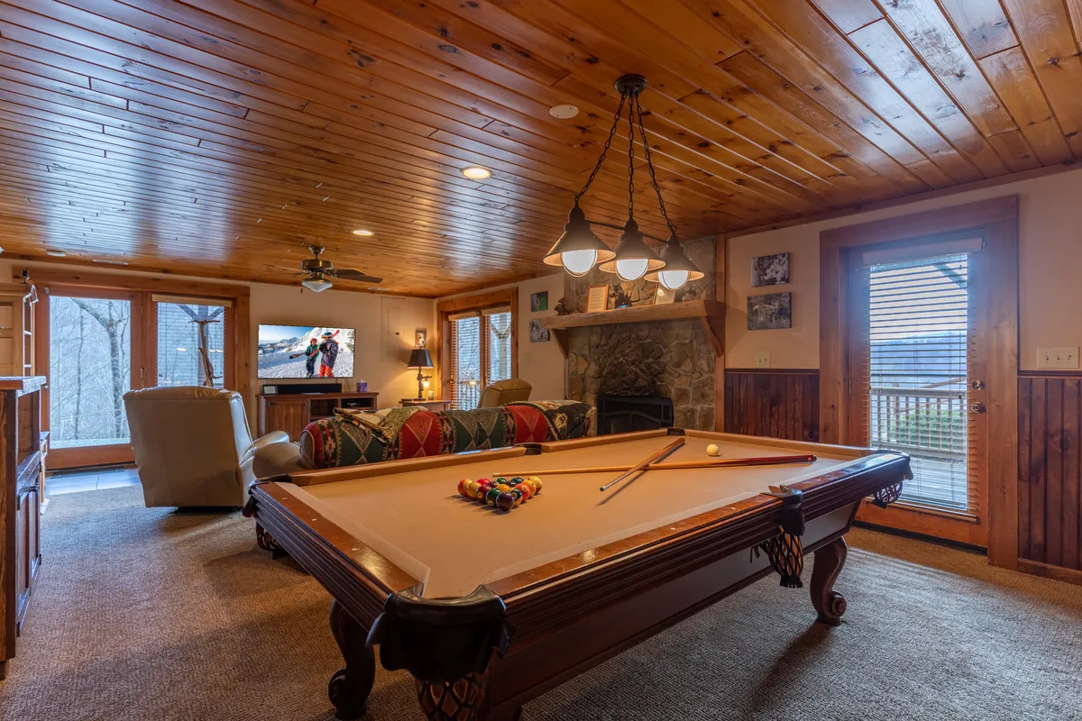 A spacious downstairs den featuring a full-size pool table centrally placed, with a crackling wood-burning fireplace on one side and a cozy sofa opposite a massive screen TV. The room is styled for gatherings, with enough space for both active games and relaxed movie watching. The ambiance is social yet inviting, with warm lighting and decorative touches that encourage fun and comfort.
